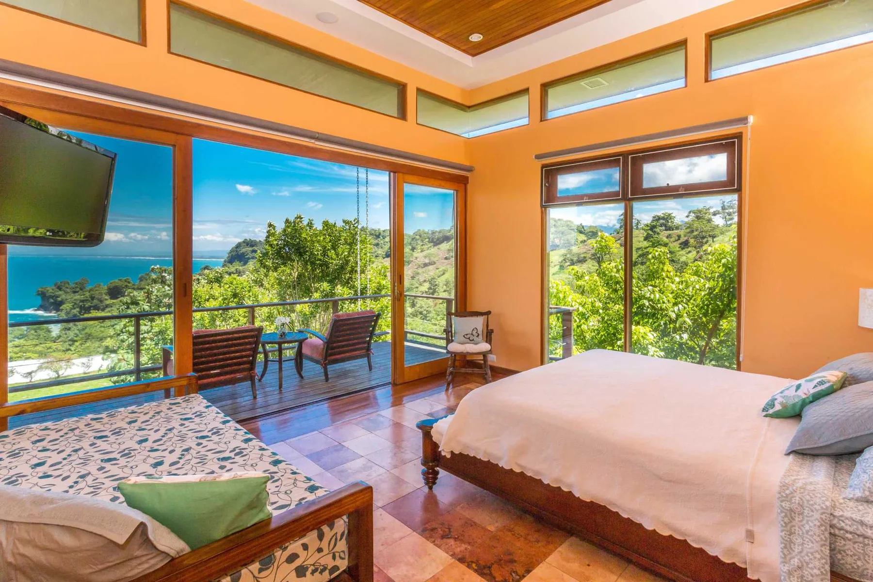 Un dormitorio luminoso tiene una cama grande, un diván estampado y puertas de vidrio que dan a un balcón con vistas al mar.