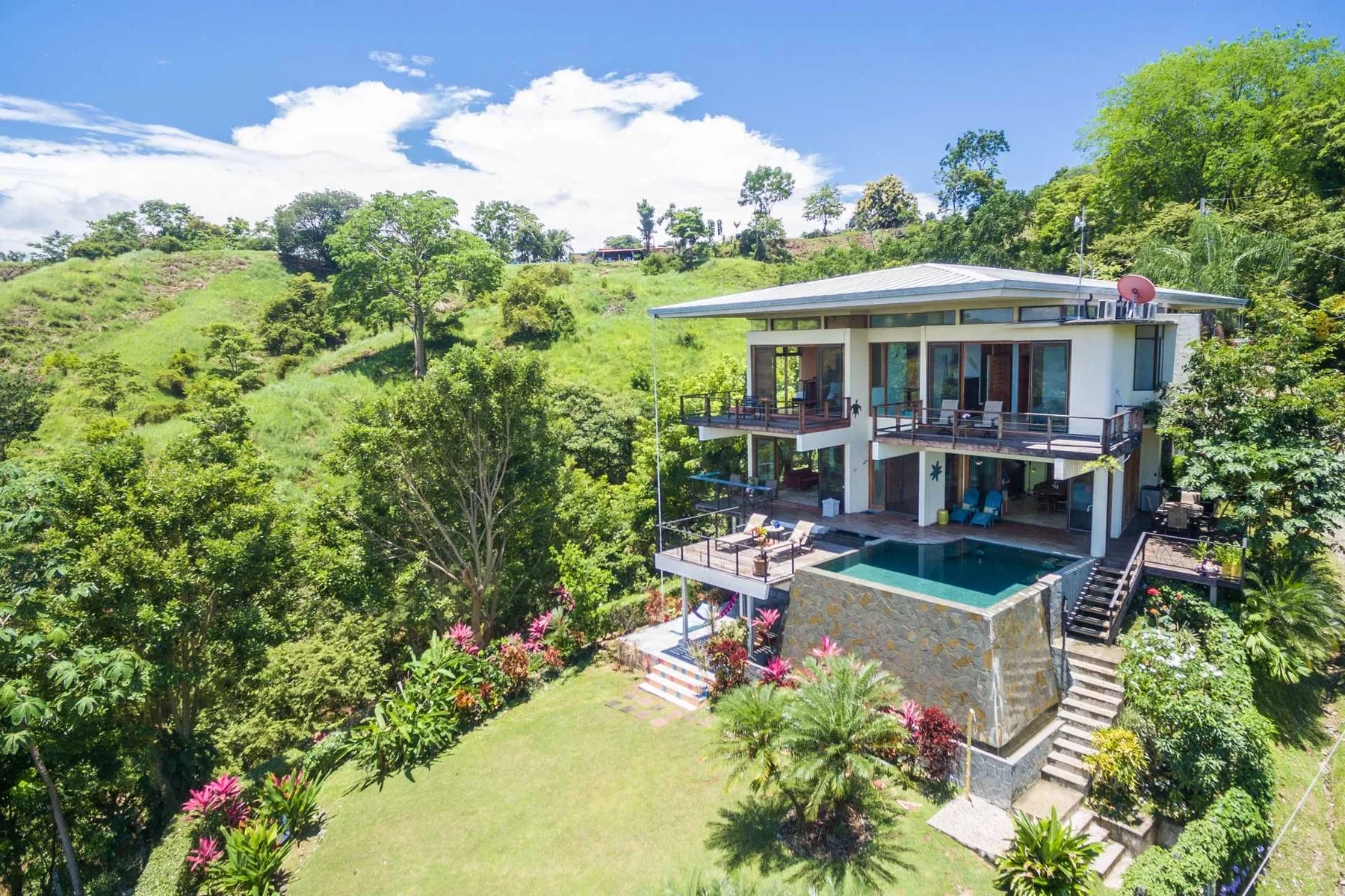 Una casa moderna de dos plantas con balcones y piscina está rodeada de colinas verdes y árboles.