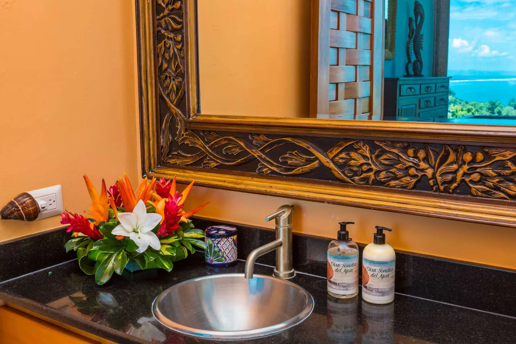 Un tocador de baño con lavabo redondo, grifo cromado, arreglo floral y espejo con marco ornamentado.