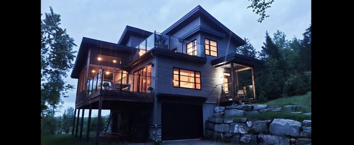 Una casa moderna entre árboles al anochecer, con ventanas iluminadas.