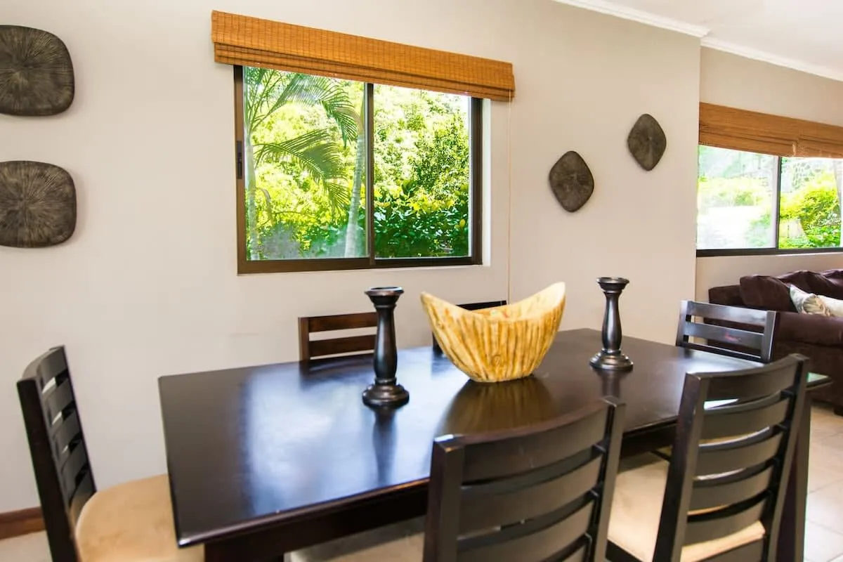 A dining area with a dark table, wooden chairs, and windows overlooking greenery.