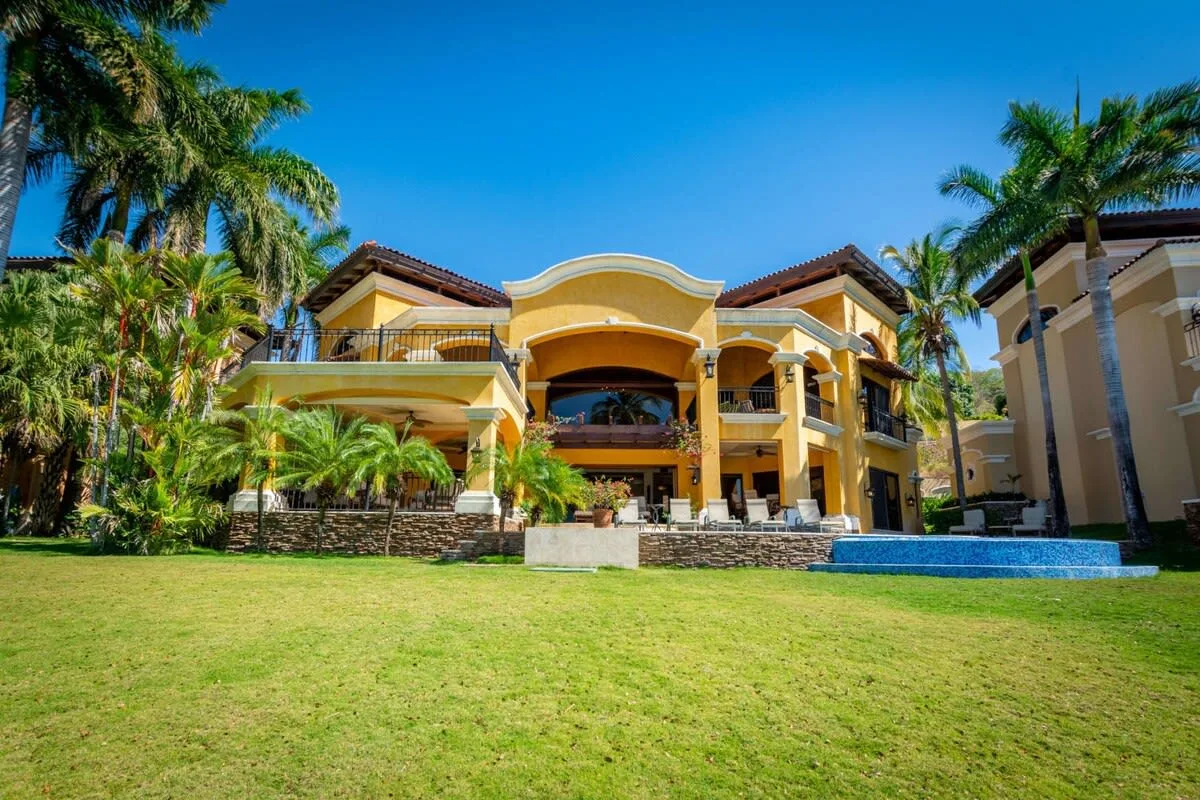 A large yellow building stands behind a green lawn, with a pool and palm trees.