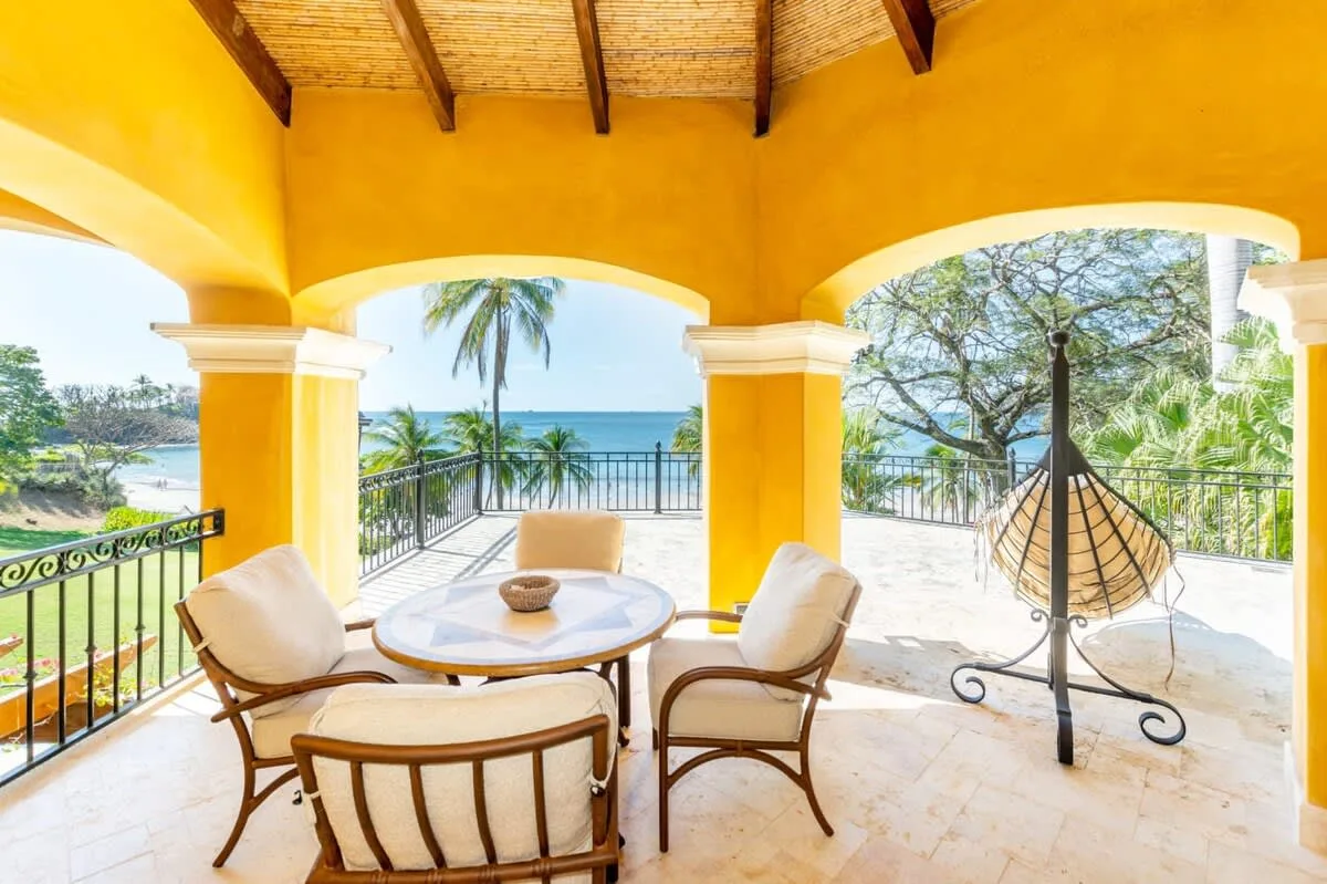 A sunny covered terrace with a round table, seating, and views of water and trees.
