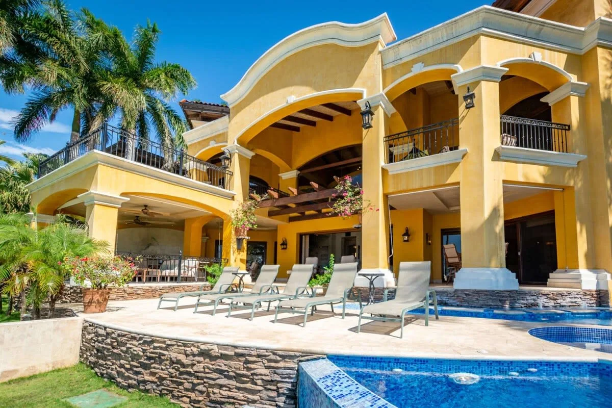 A large yellow house overlooks a swimming pool and patio with lounge seating.
