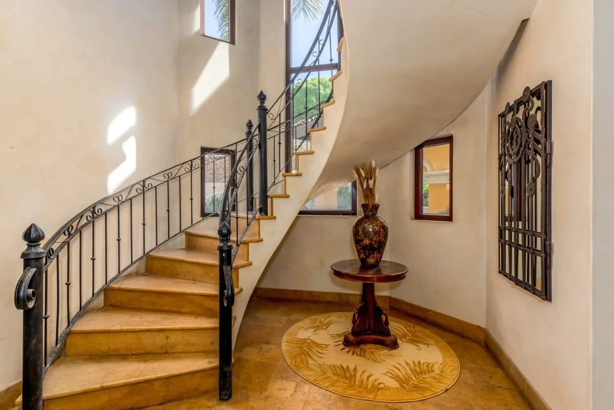 A curved staircase with a dark railing rises beside a small table and decorative vase.