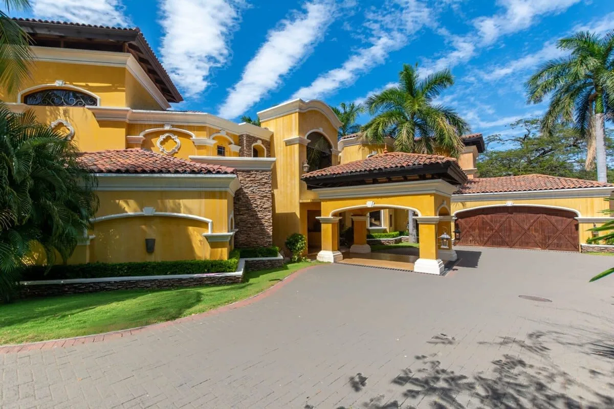 A large yellow house with a covered driveway and palm trees.