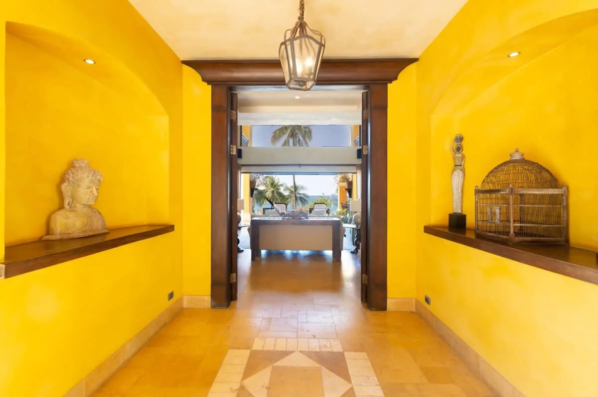 A bright yellow hallway leads through a dark wood doorway to a sunlit seating area.