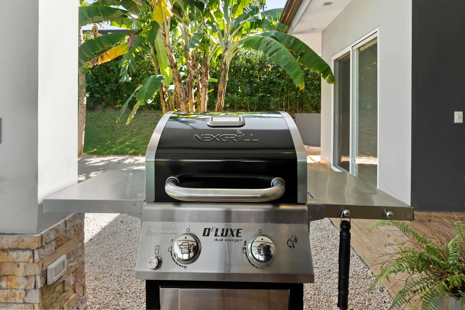An outdoor grill stands on a patio beside a glass door, with leafy plants behind it.