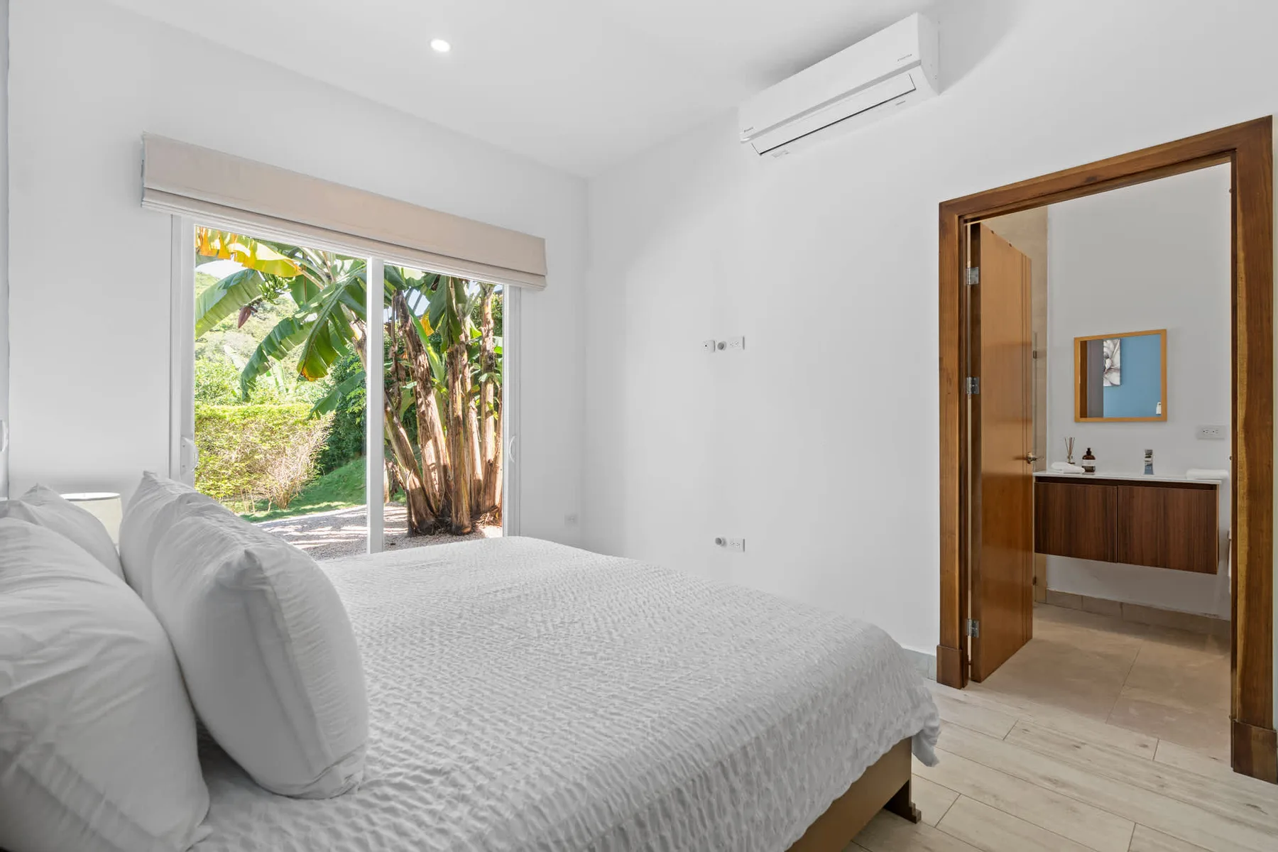 A bright bedroom with a bed beside a large window overlooking greenery.