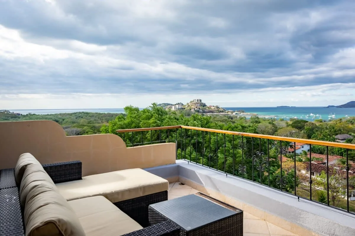 A terrace seating area overlooks green hills and the sea beneath a cloudy sky.