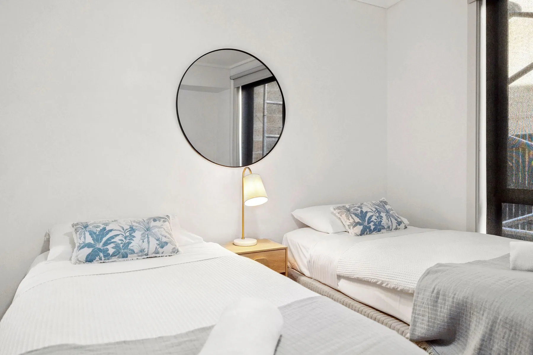 A bright bedroom with two beds and a round wall mirror.