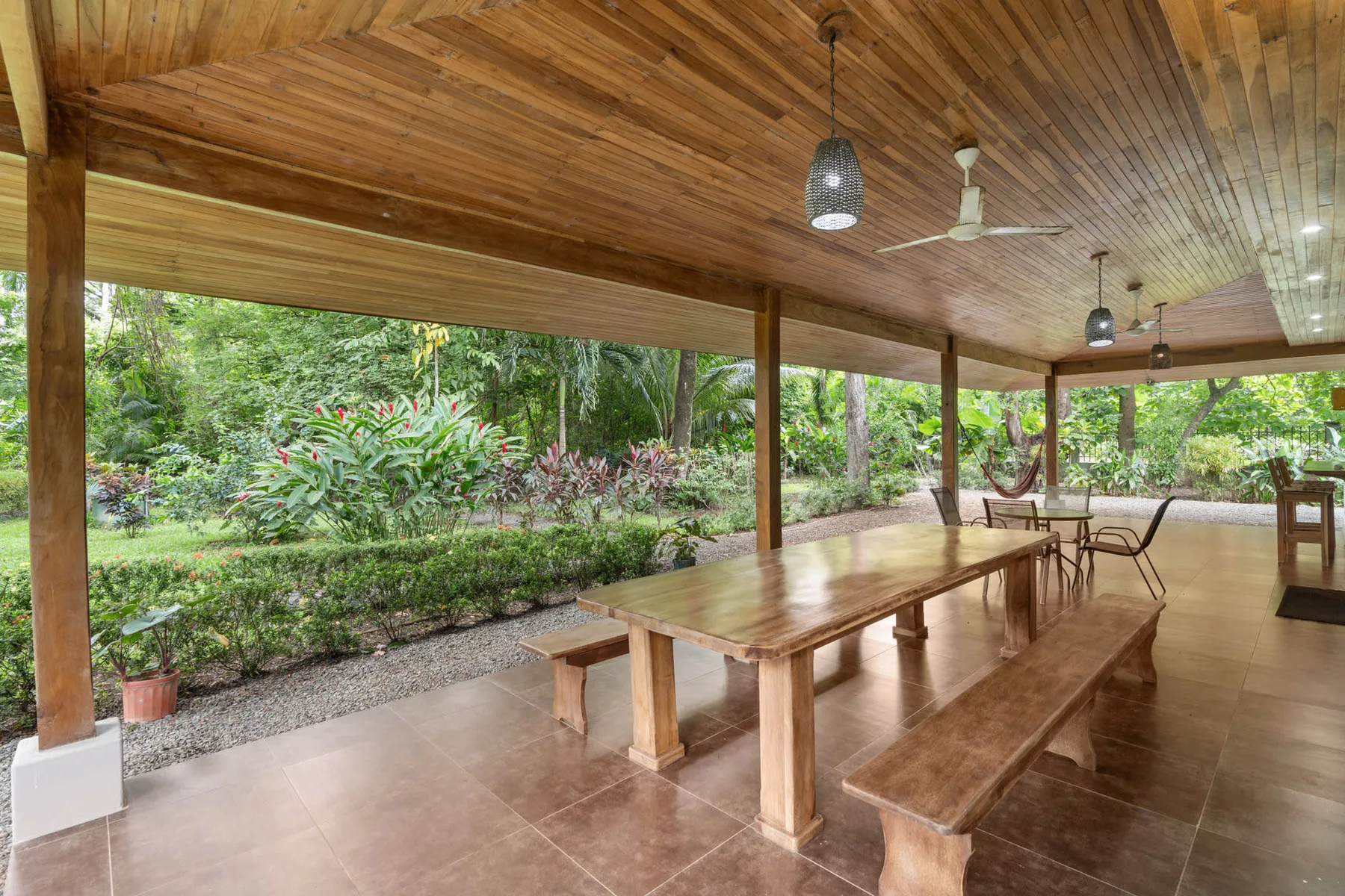 A covered outdoor dining area with wooden tables and benches beside a garden.