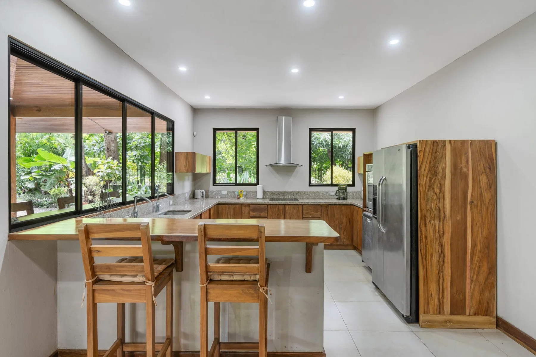 A modern kitchen with a long counter, wooden stools, and large windows.
