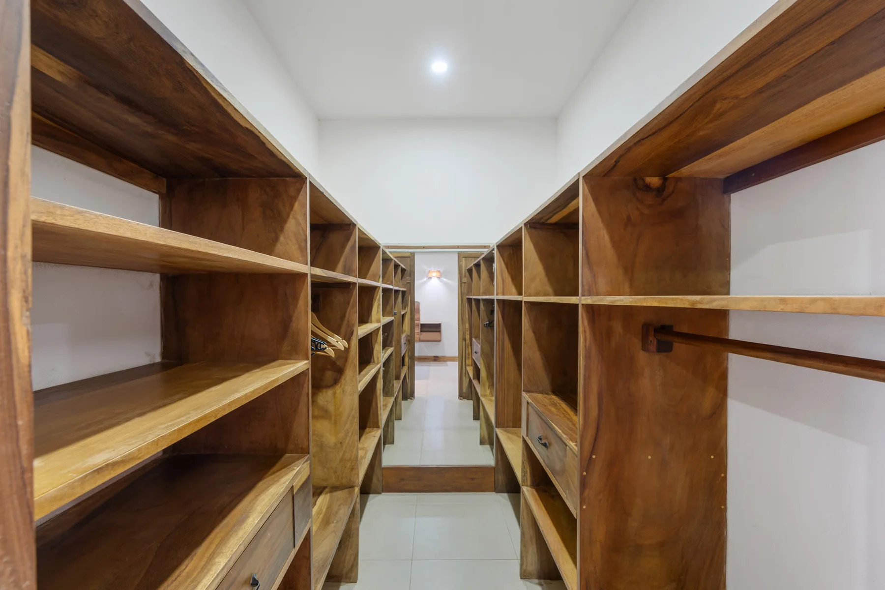 A narrow storage room has wooden shelving along both sides of a tiled aisle.