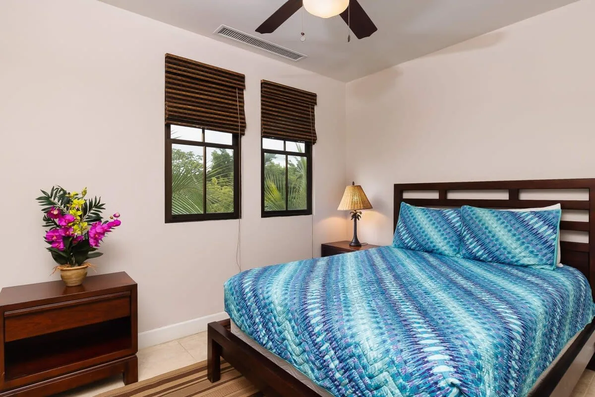 Un dormitorio con una cama vestida con ropa de cama azul estampada y dos ventanas.