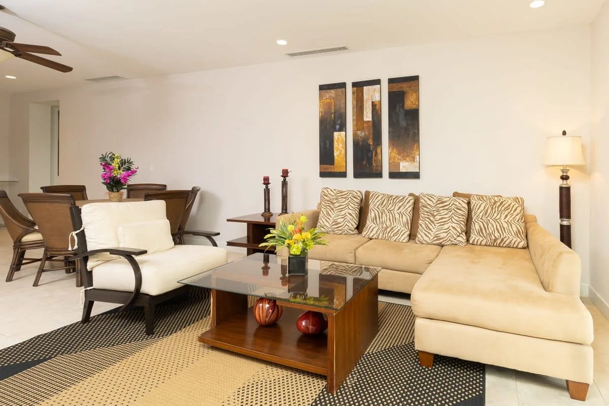 Una sala de estar con asientos beige, una mesa de centro de vidrio y arte en la pared.