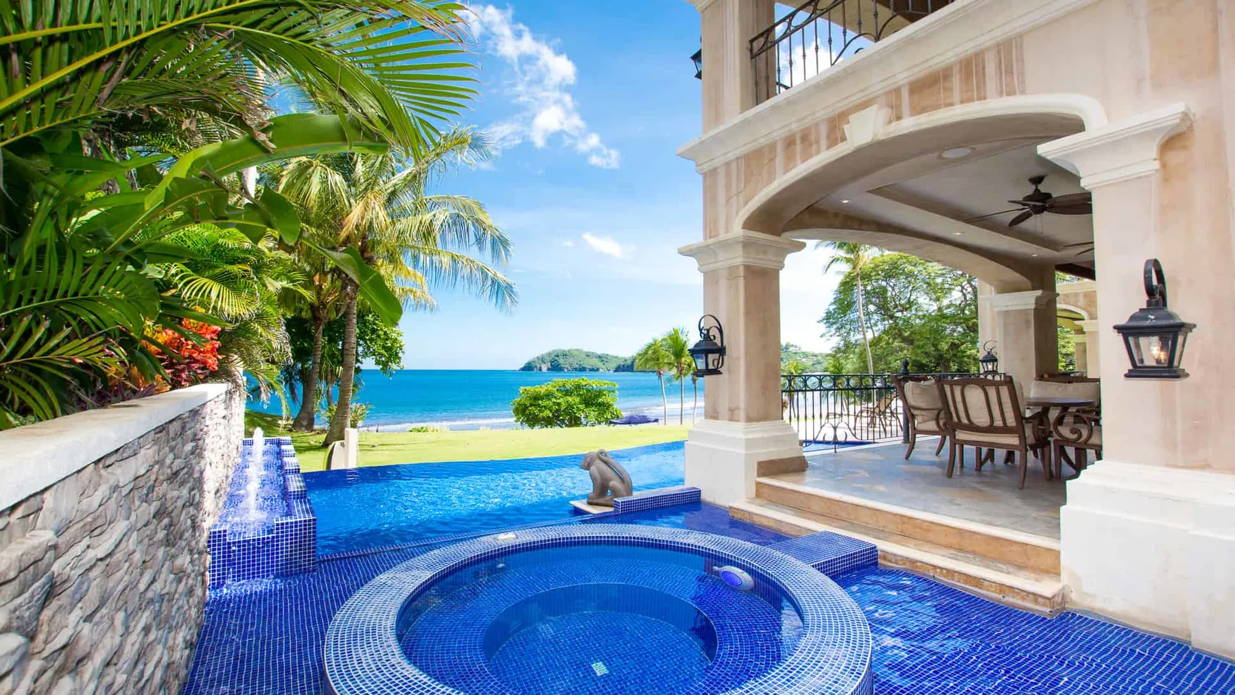A tiled pool and round spa sit beside a covered patio overlooking the sea.