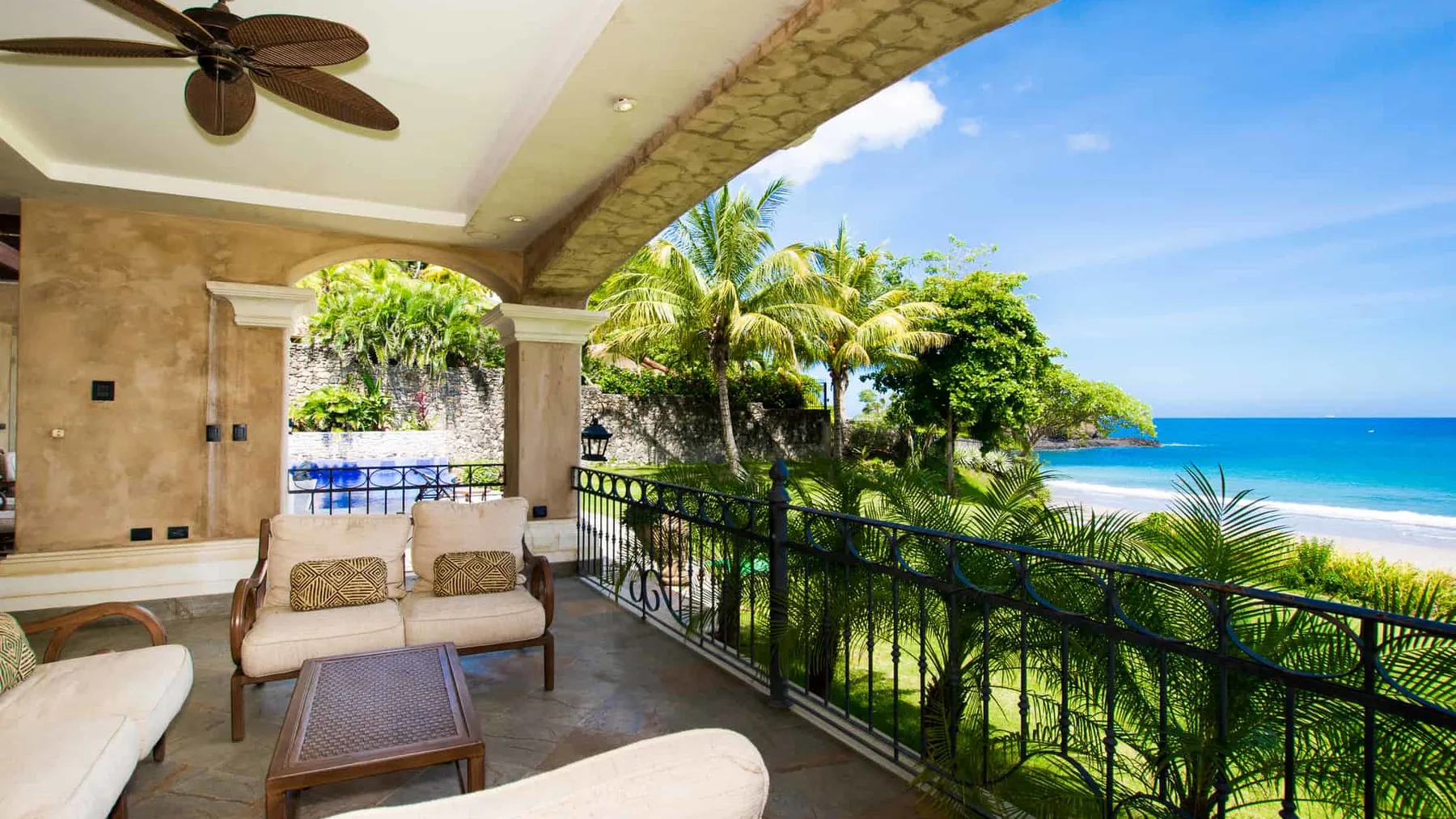 A covered patio with seating overlooks tropical greenery and the ocean.