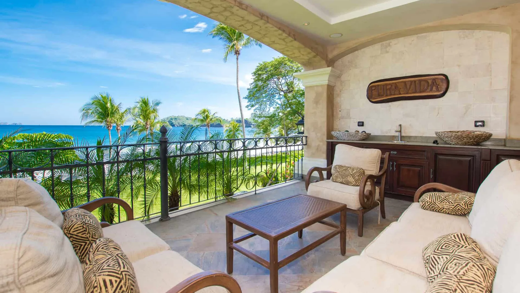 A covered terrace with cushioned seating overlooks the ocean and palm trees.
