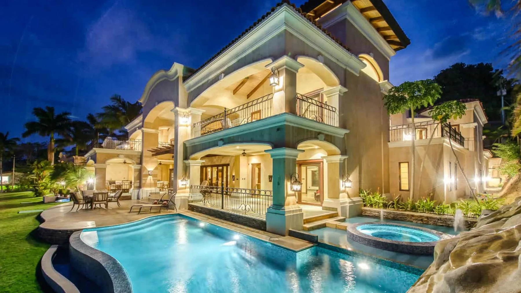 A large illuminated villa stands beside a swimming pool at night.