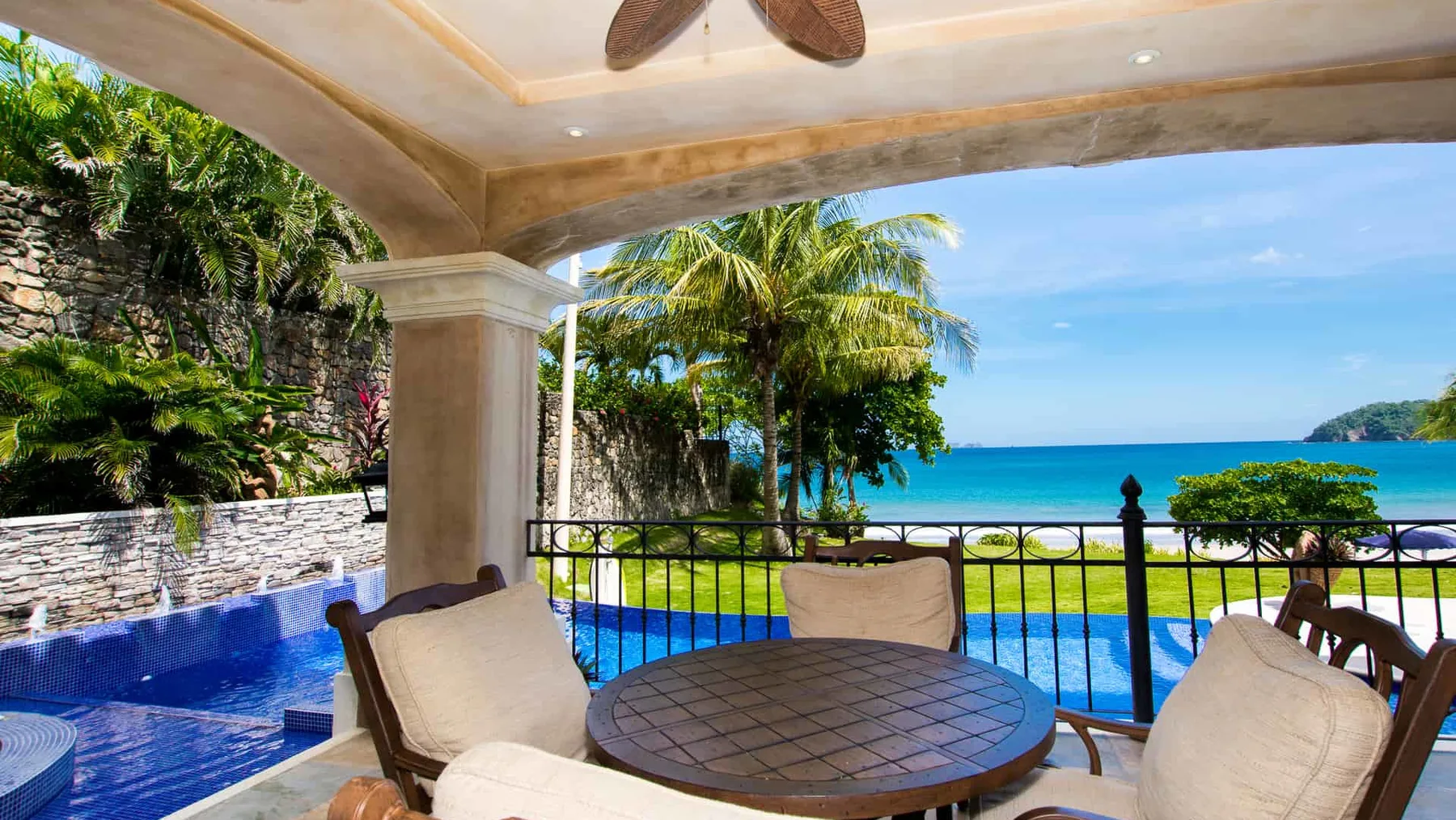 A covered patio with seating overlooks a pool and the ocean.