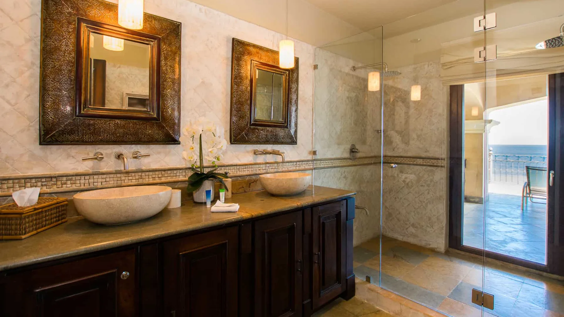 A bathroom vanity with two sinks and a glass shower beside a bright doorway.