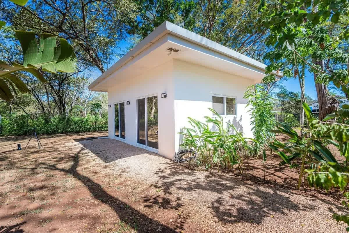 A small white building stands in a leafy outdoor setting.