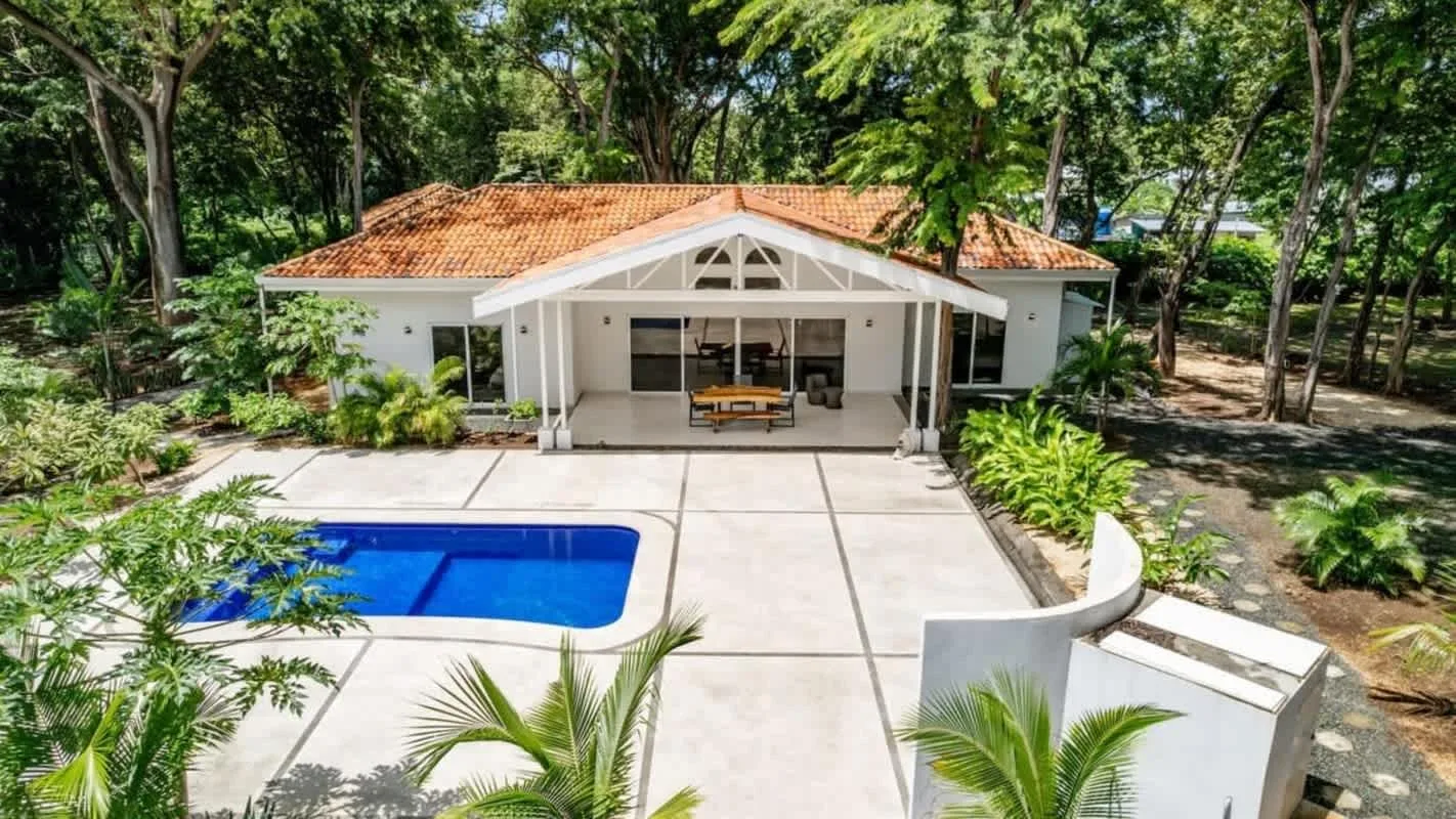 A white house opens onto a paved patio with a blue swimming pool, surrounded by trees.
