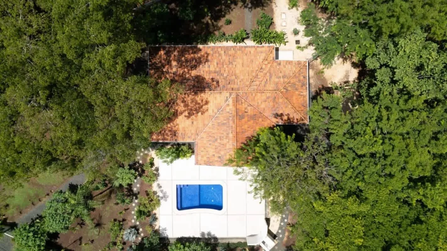 Aerial view of a tiled-roof house surrounded by trees, with a swimming pool nearby.