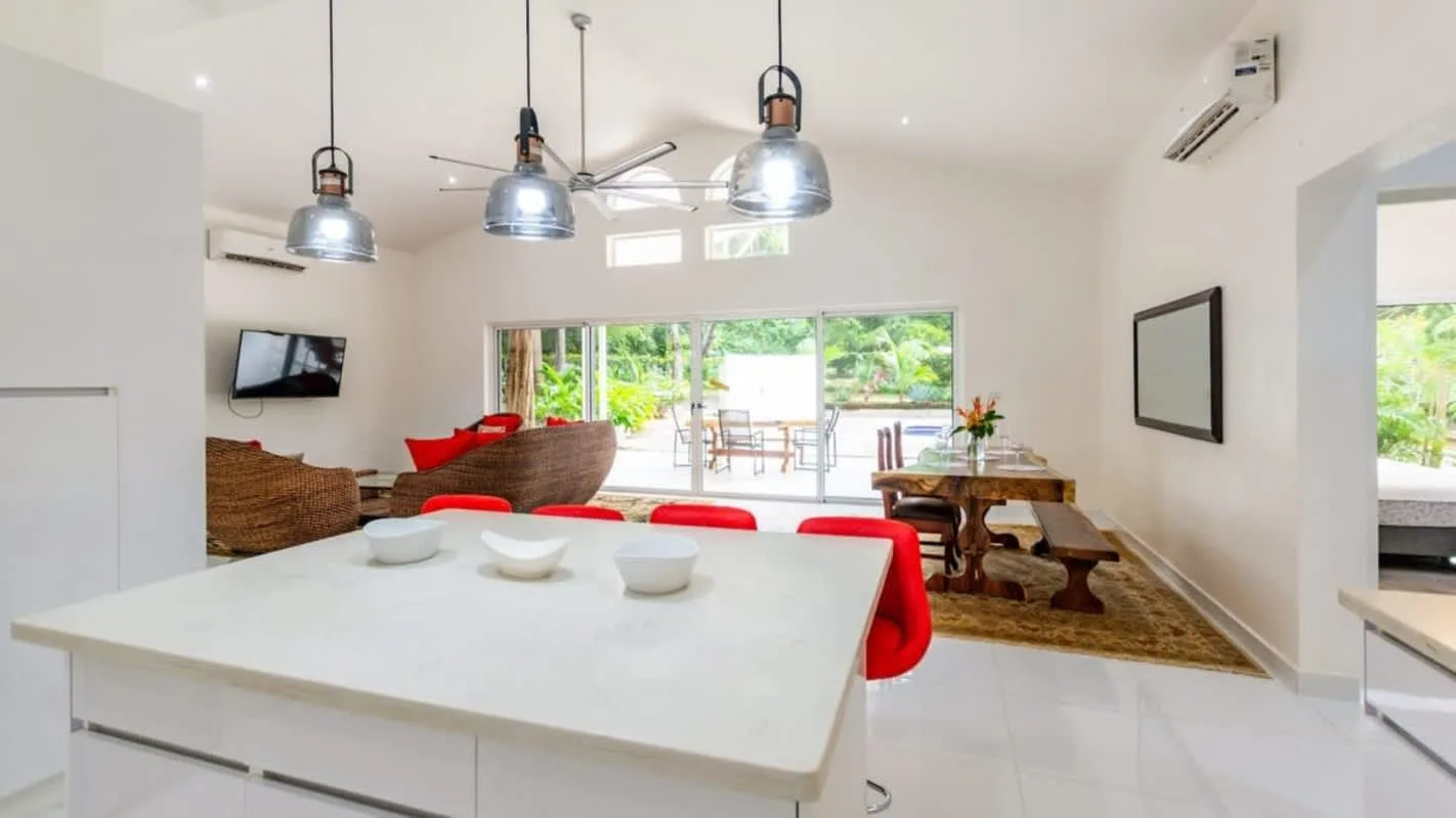 A bright open-plan dining and living area has a large white island and windows overlooking greenery.