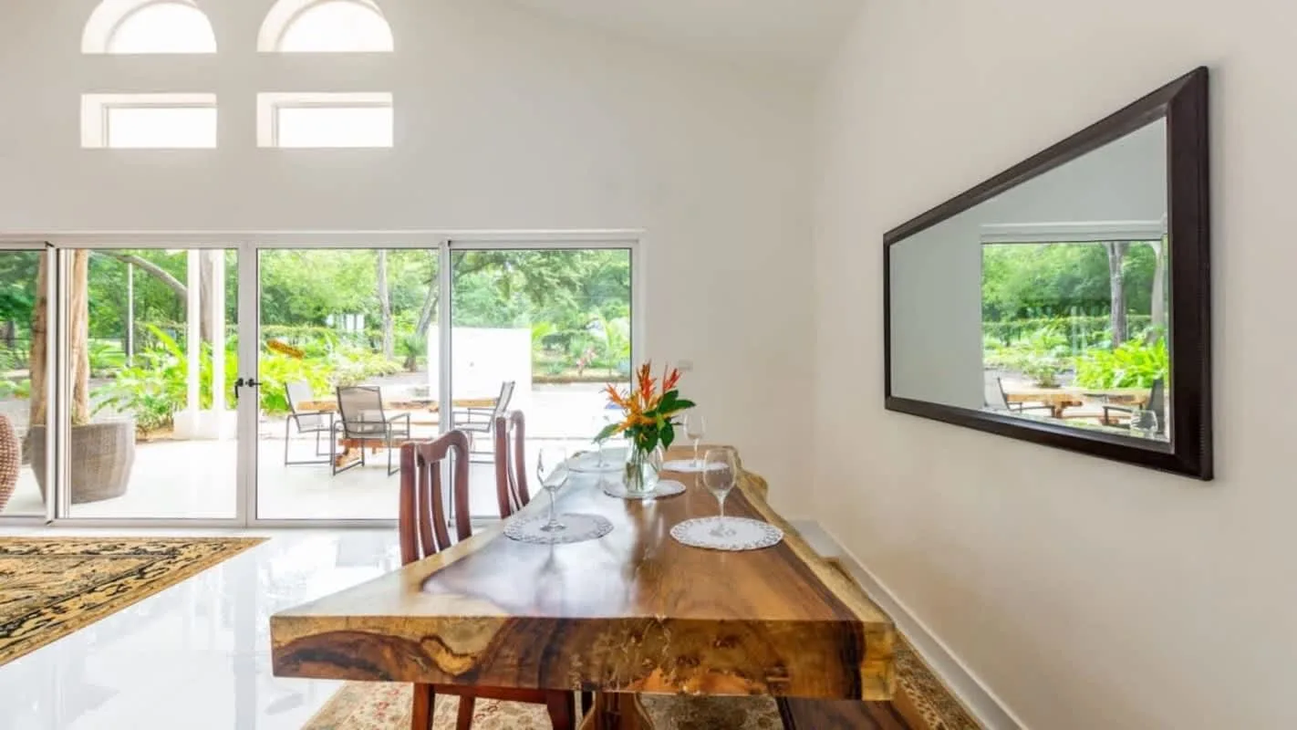 A bright dining area with a wooden table beside large windows overlooking greenery.