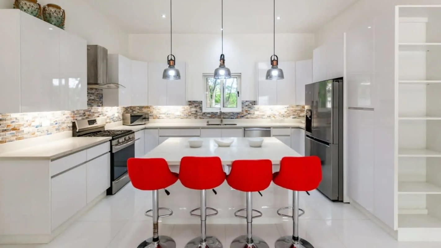 A bright modern kitchen has white cabinets, a long island, and red bar stools.