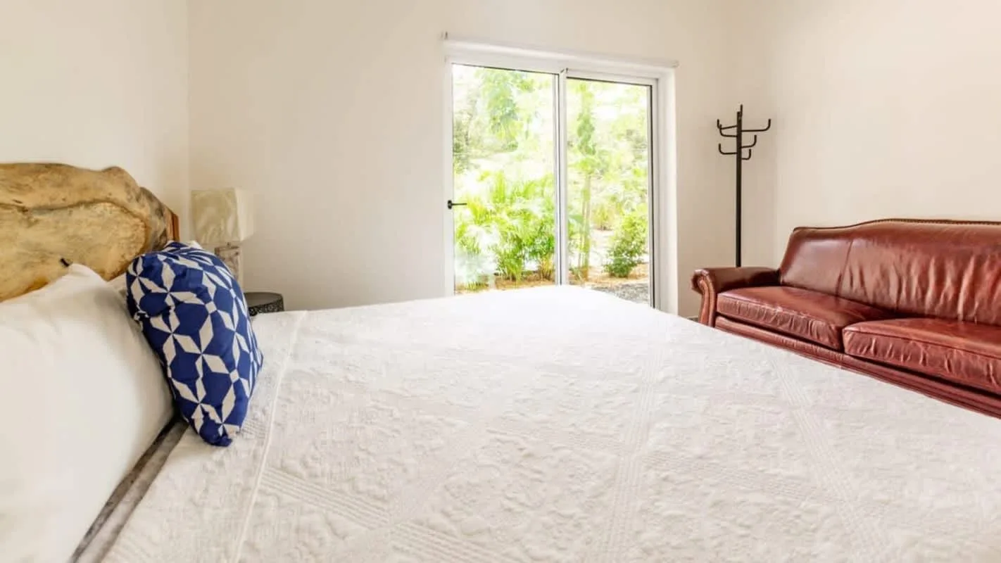 A bedroom with a white bed, blue patterned pillow, and brown seating beside a glass door.