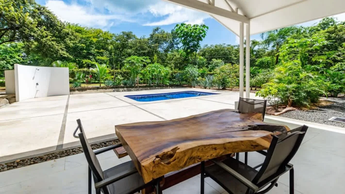 A covered outdoor dining area overlooks a swimming pool and leafy garden.