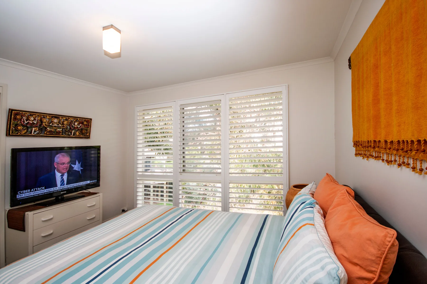 A bedroom with a striped bed, orange pillows, and a television beside shuttered windows.