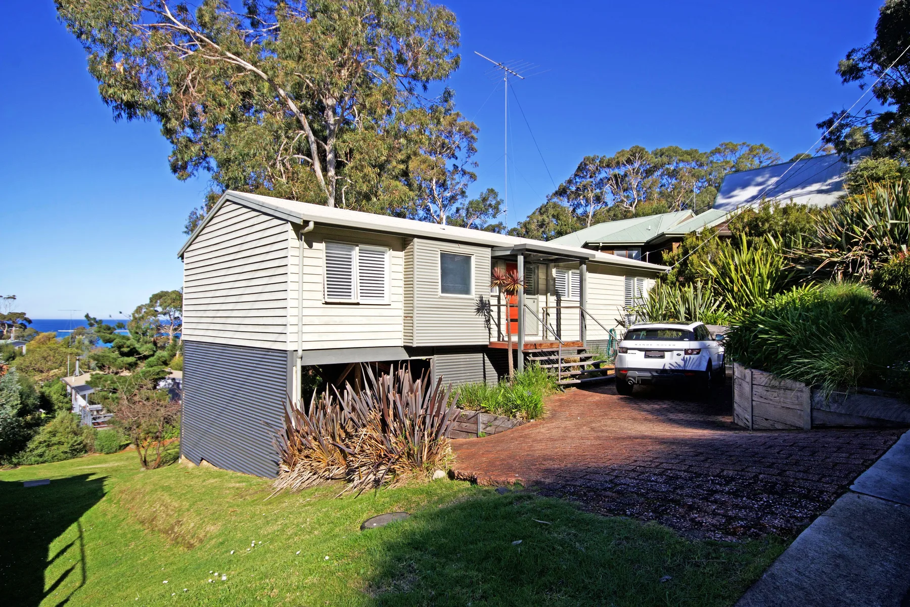 A raised house beside a sloping driveway, with trees and greenery around it.
