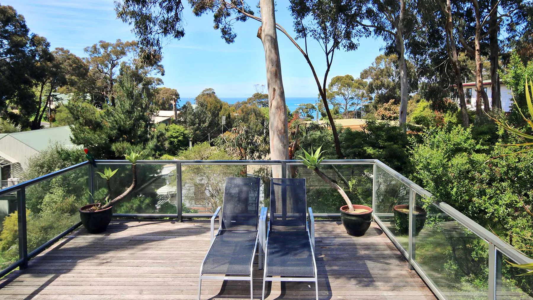A wooden deck with glass railings overlooks trees and a glimpse of the water.