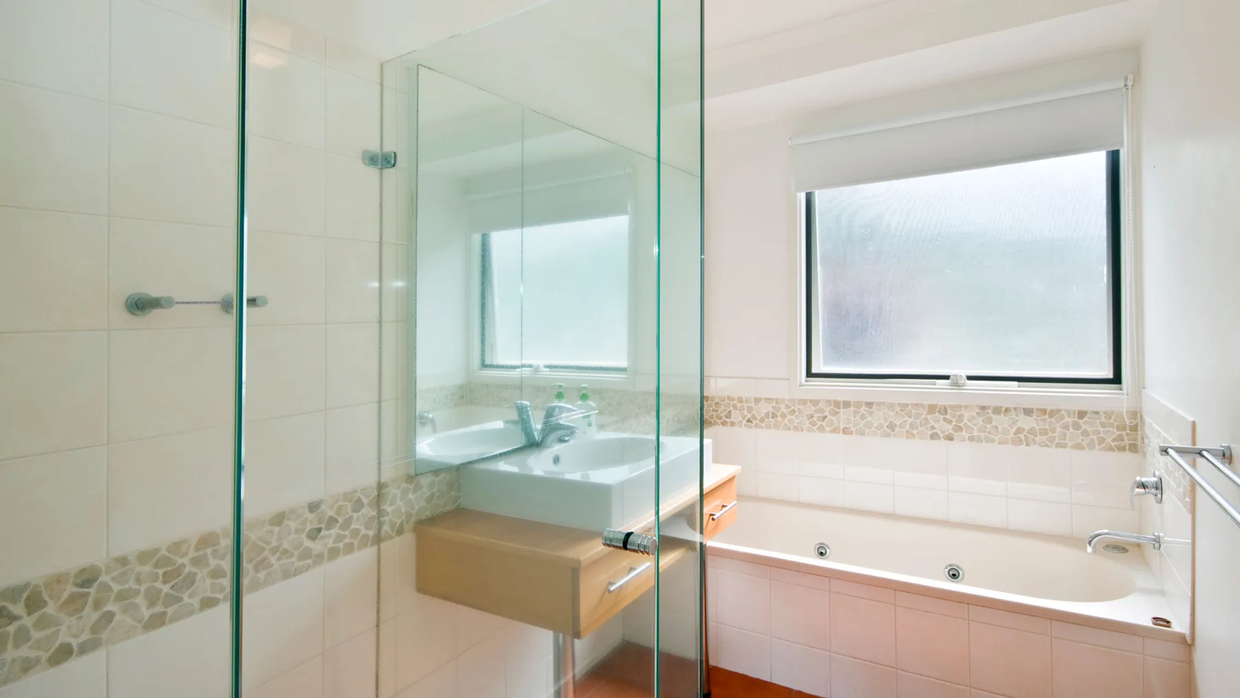 A bright bathroom with a glass shower, sink, and bathtub beneath a window.