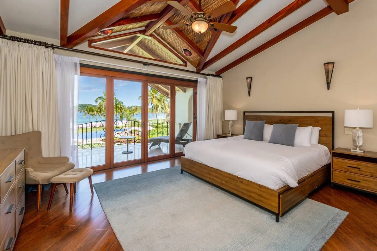A bedroom with a large bed, wooden ceiling beams, and glass doors opening to a balcony.