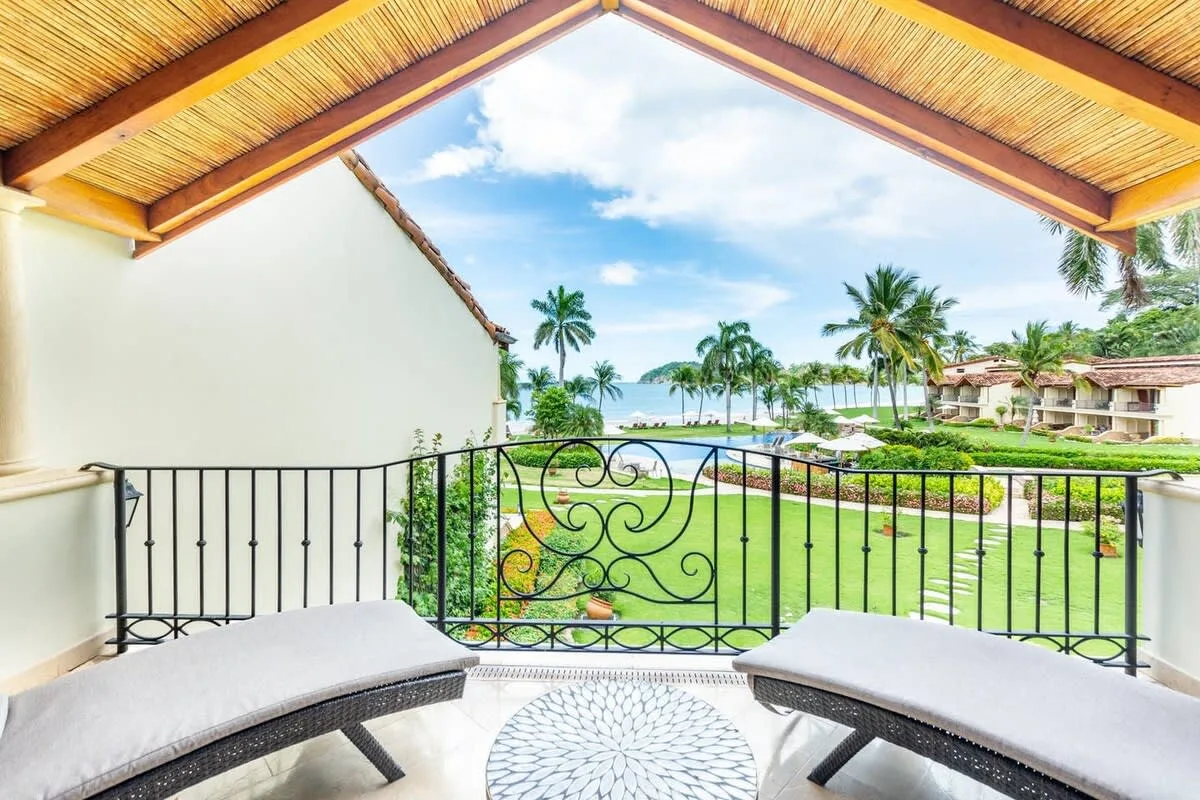 A covered balcony overlooks a garden, palm trees, and the ocean.