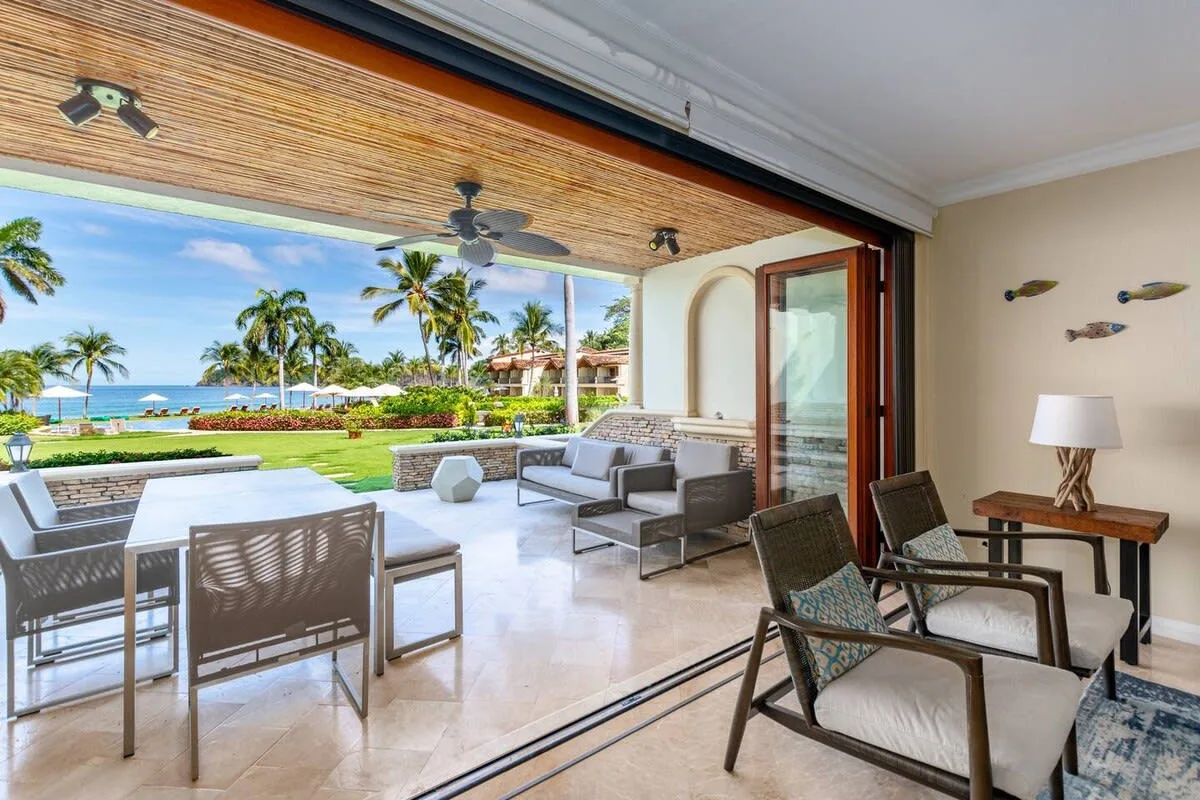 A covered patio with seating opens onto a tropical garden and ocean view.
