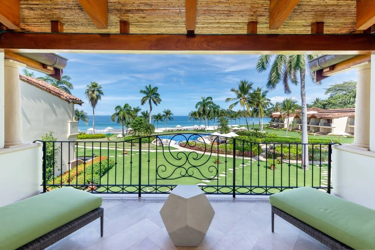 A covered balcony overlooks a garden, palm trees, and the ocean.
