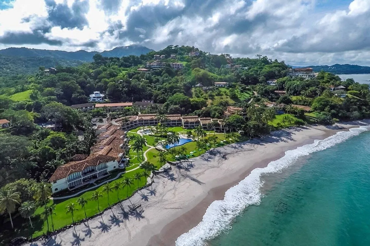 Shared amenity: Aerial view of a beachfront property beside a sandy shore and green hills.