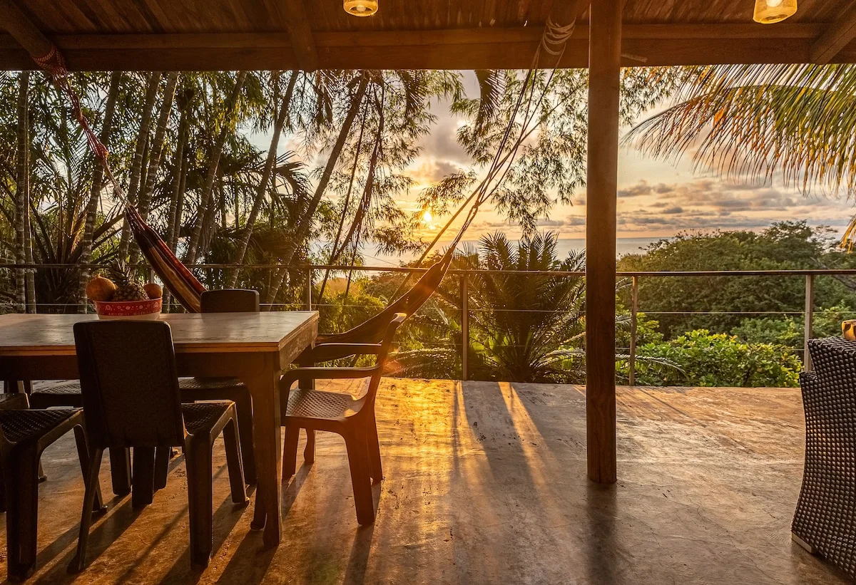 Una zona cubierta de comedor al aire libre, con mesa, asientos y hamaca, da a vegetación tropical al atardecer.