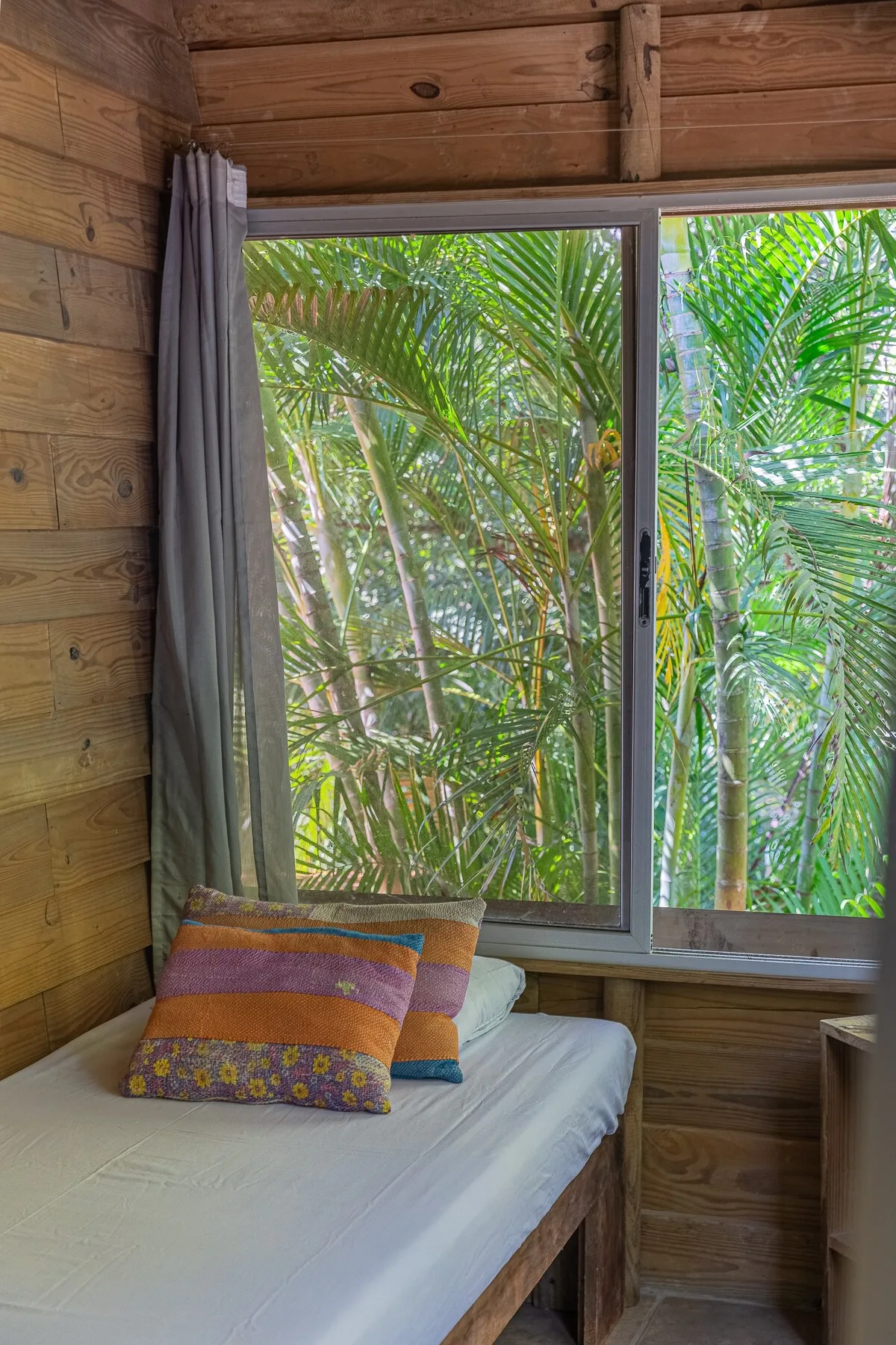 Dormitorio de madera con una cama junto a una ventana grande que da a palmeras.