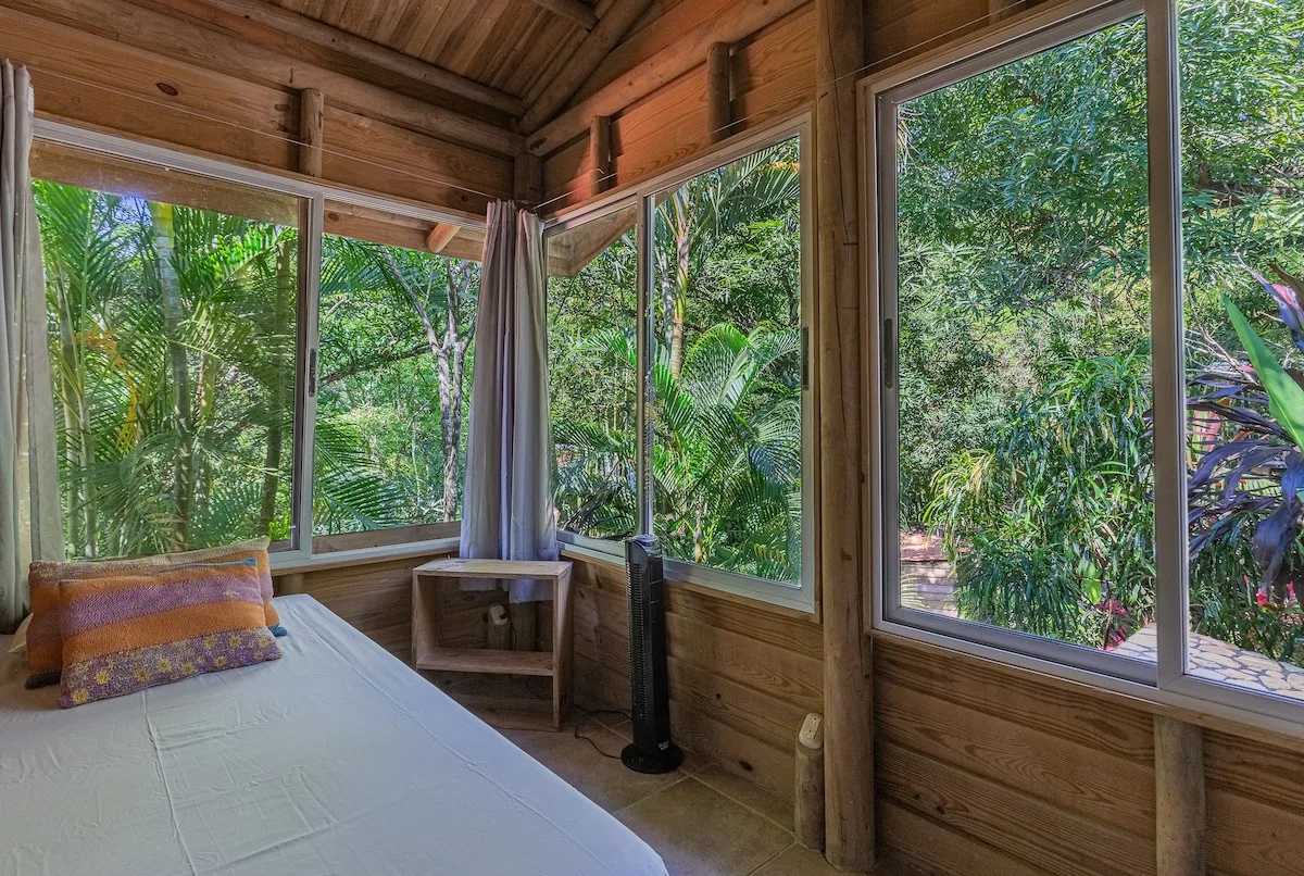 Un dormitorio de madera con una cama junto a grandes ventanas que dan a una exuberante vegetación.