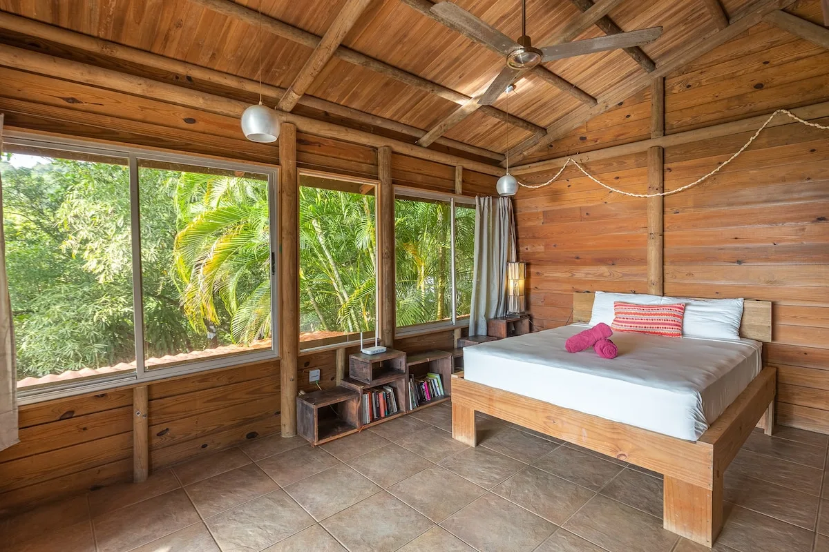 Un dormitorio de madera con una cama junto a amplias ventanas con vistas a la vegetación.