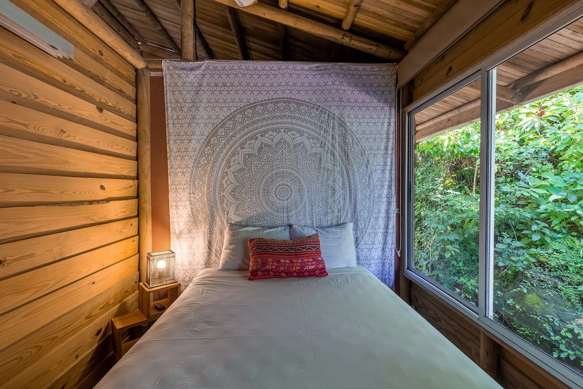 Dormitorio con una cama, un tapiz estampado y grandes ventanas con vistas a la vegetación.