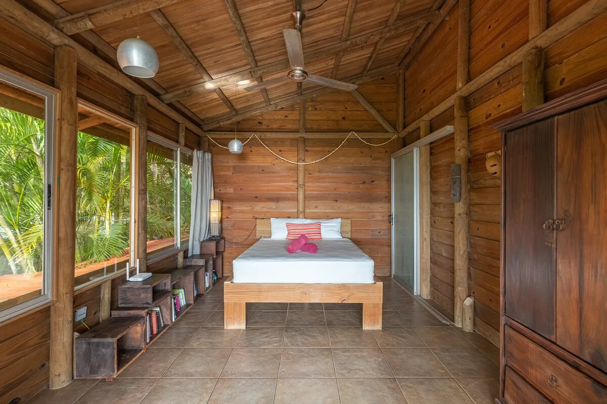 Un dormitorio de madera con una cama entre amplias ventanas y un armario oscuro.