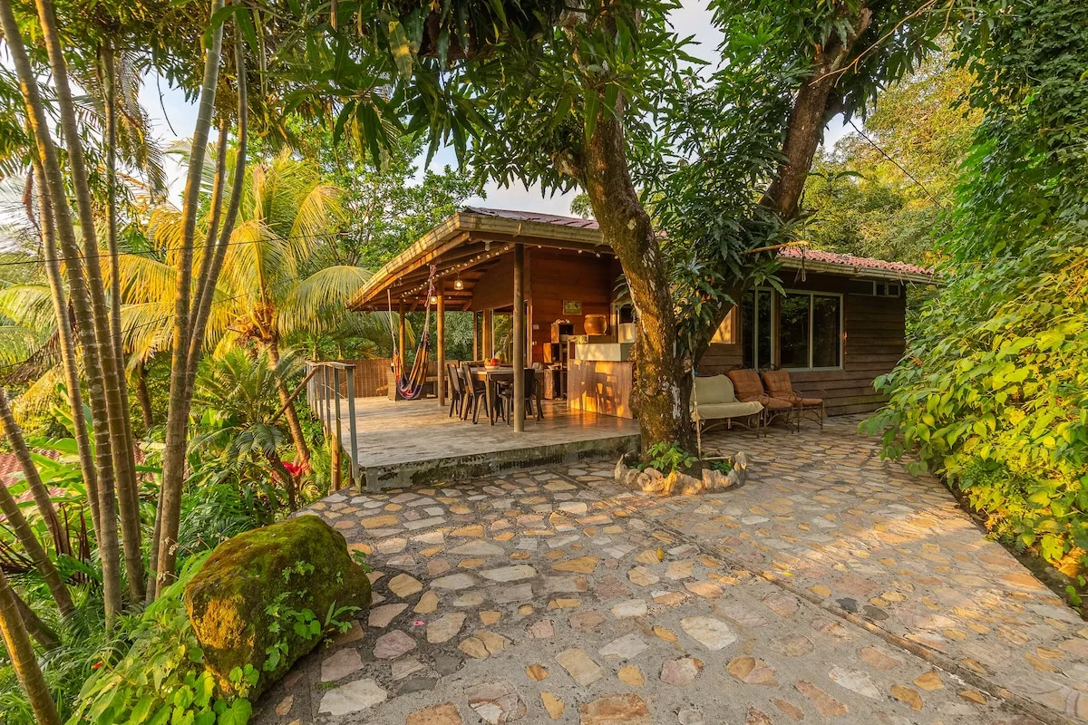 Una cabaña de madera y una zona de estar cubierta al aire libre están rodeadas de vegetación tropical.